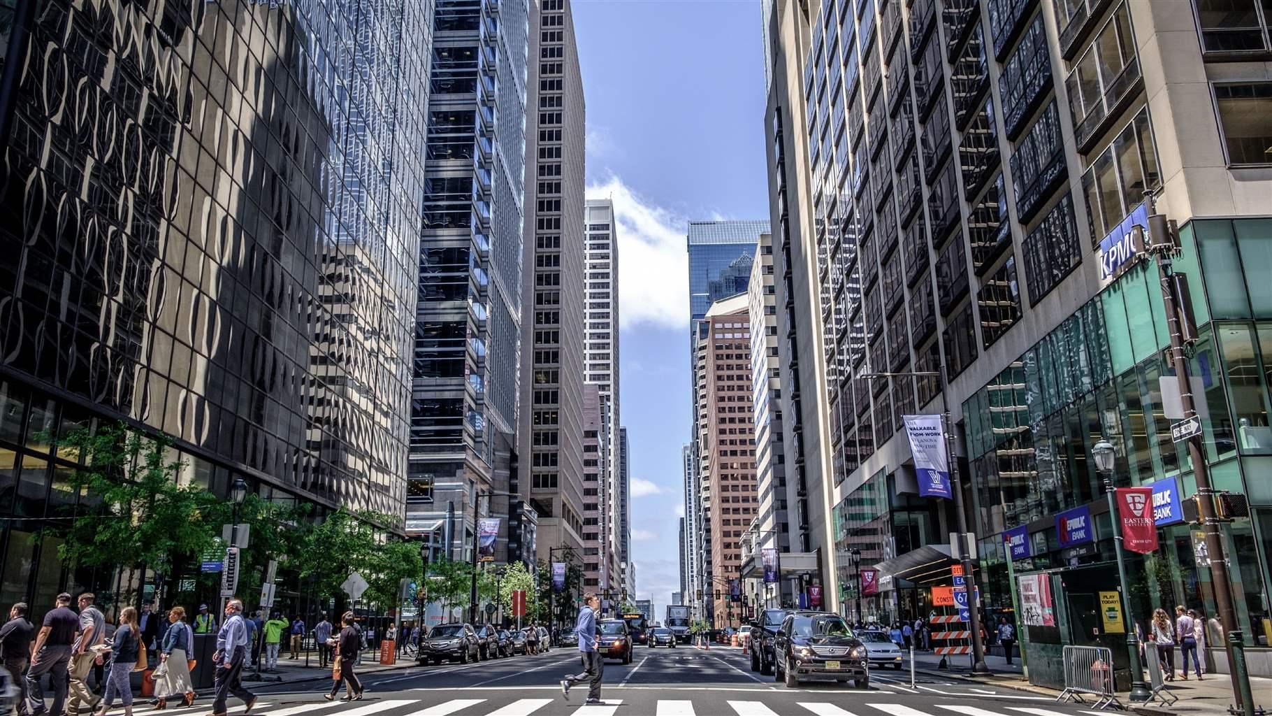 Tall office buildings line both sides of Market Street in Center City Philadelphia. Cars wait at the intersection of 16th and Market streets while pedestrians cross on a clear, sunny day. 