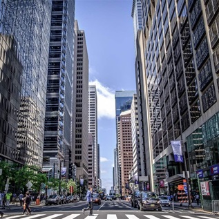 Tall office buildings line both sides of Market Street in Center City Philadelphia. Cars wait at the intersection of 16th and Market streets while pedestrians cross on a clear, sunny day. 