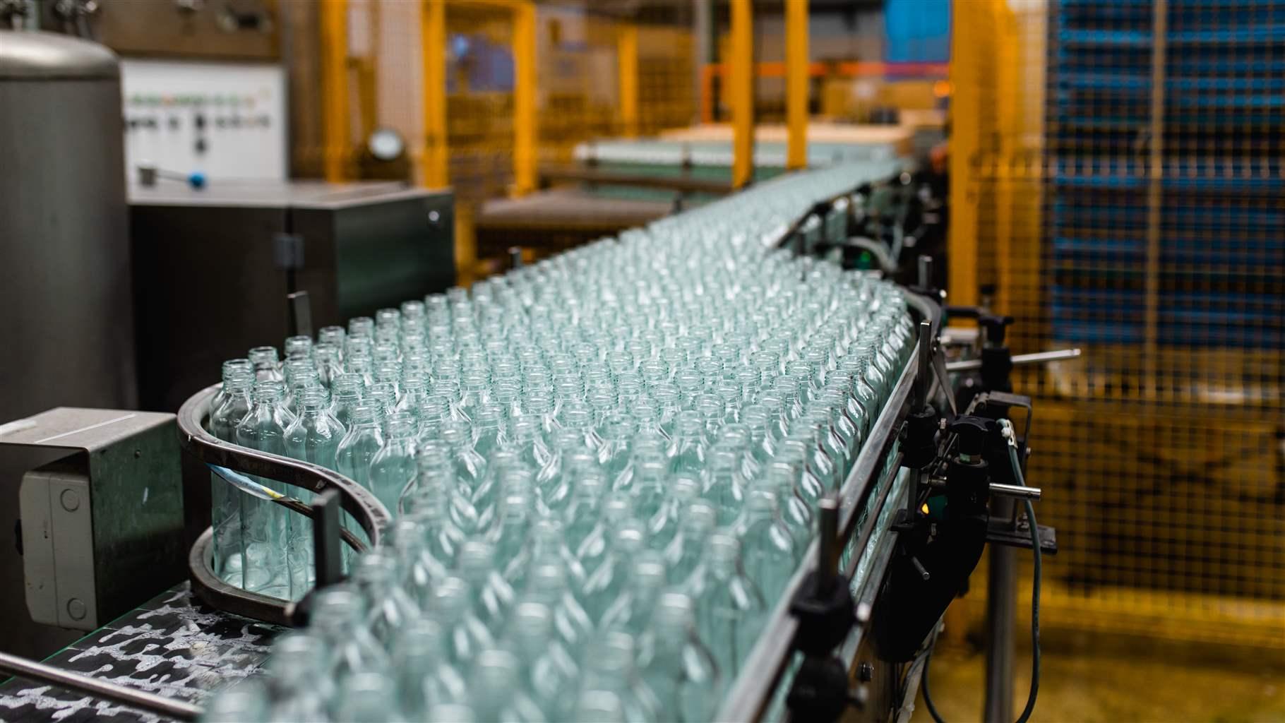  Hundreds of clear bottles, held in place by black rails, are crammed onto a conveyor belt moving through a factory.  