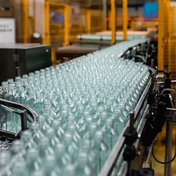  Hundreds of clear bottles, held in place by black rails, are crammed onto a conveyor belt moving through a factory.  
