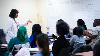 An instructor in a white medical coat lectures to a classroom of nursing students in front of a white board and model of a skeleton.