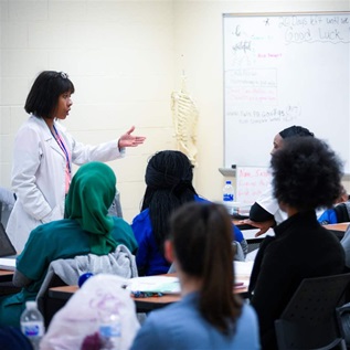 An instructor in a white medical coat lectures to a classroom of nursing students in front of a white board and model of a skeleton.