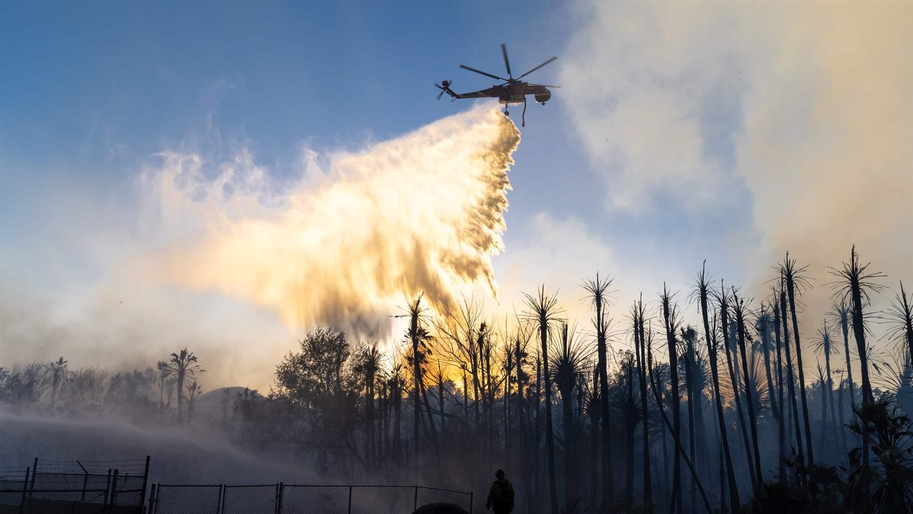   A helicopter drops a large plume of water on a smoldering wildfire, with scorched trees, some chain-link fencing, and the silhouette of a firefighter visible in the foreground. The plume is refracting light from the sun, which sits behind a small mountain on the horizon beneath a cloudless sky.