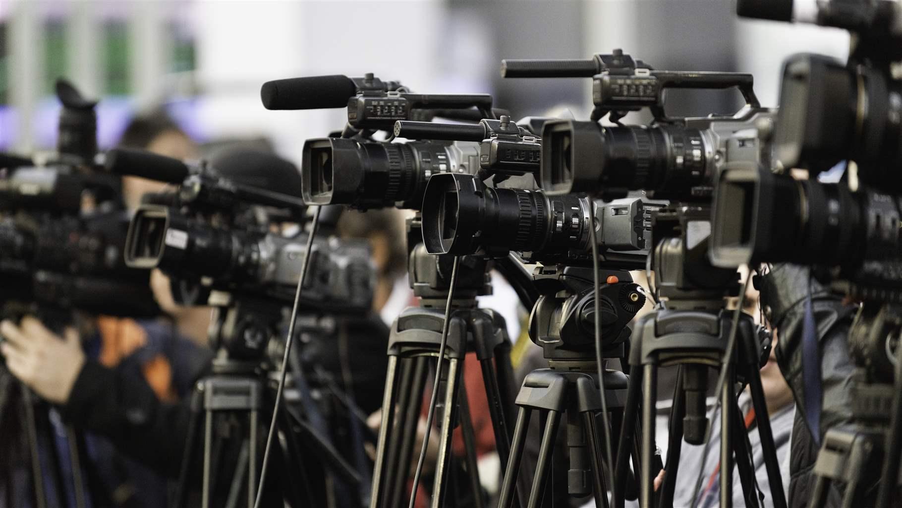 Five video cameras on tripods stand in a line, with one reporter holding a camera slightly out of focus, all aimed at the same subject.
