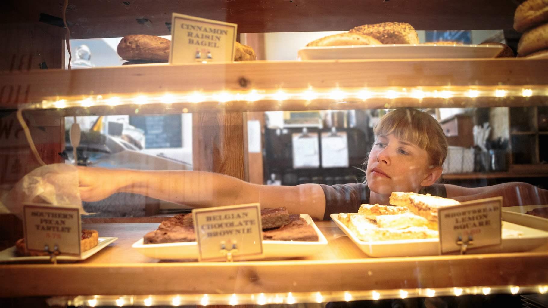 A bakery shop employee with brown hair and wearing a gray shirt reaches into a lighted pastry case where the store’s array of baked goods are displayed.
