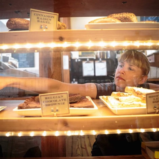 A bakery shop employee with brown hair and wearing a gray shirt reaches into a lighted pastry case where the store’s array of baked goods are displayed.