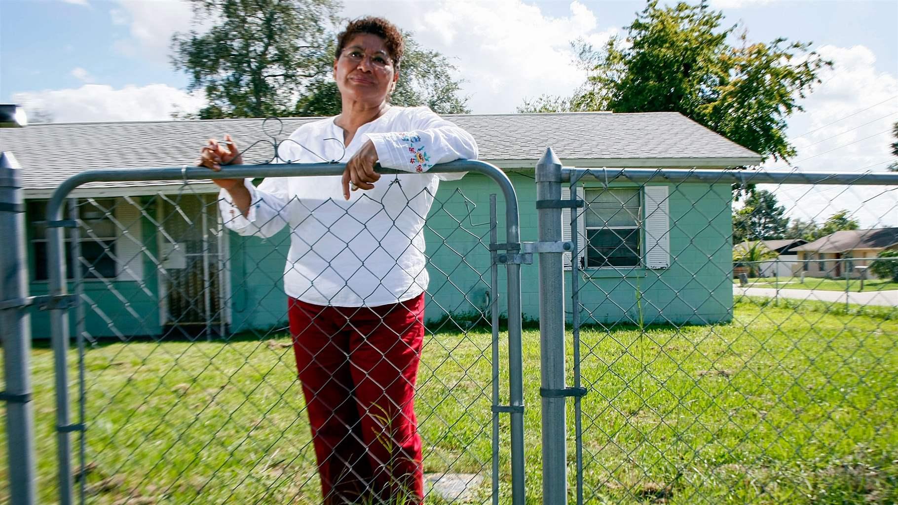 A woman stands at her chain link fence in front of her light green manufactured home. 