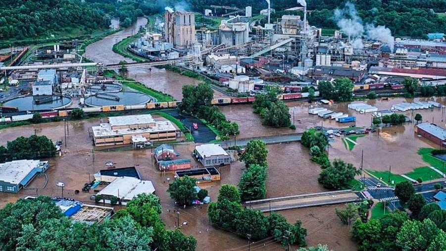 An aerial photo shows a section of a small city with industrial buildings, warehouses, and a train on tracks that run over a river. That river is swollen with brown floodwater that also fills most of the city streets and parks. 