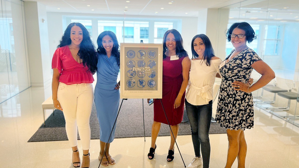 Five women stand smiling at the photographer around a framed standing poster marking The Pew Charitable Trusts’ 75th anniversary.