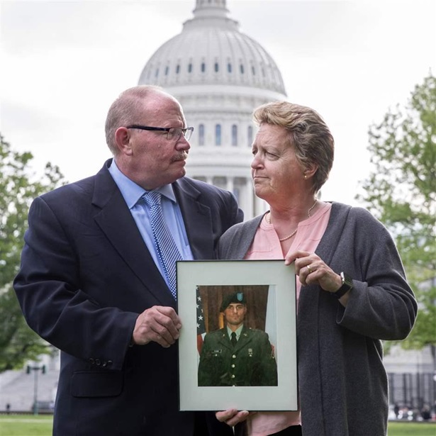 A white couple with gray hair looks at each other while holding a picture of a young man in a military uniform. The U.S. Capitol building is behind them. Photo provided by the Partnership to Fight Infectious Disease.