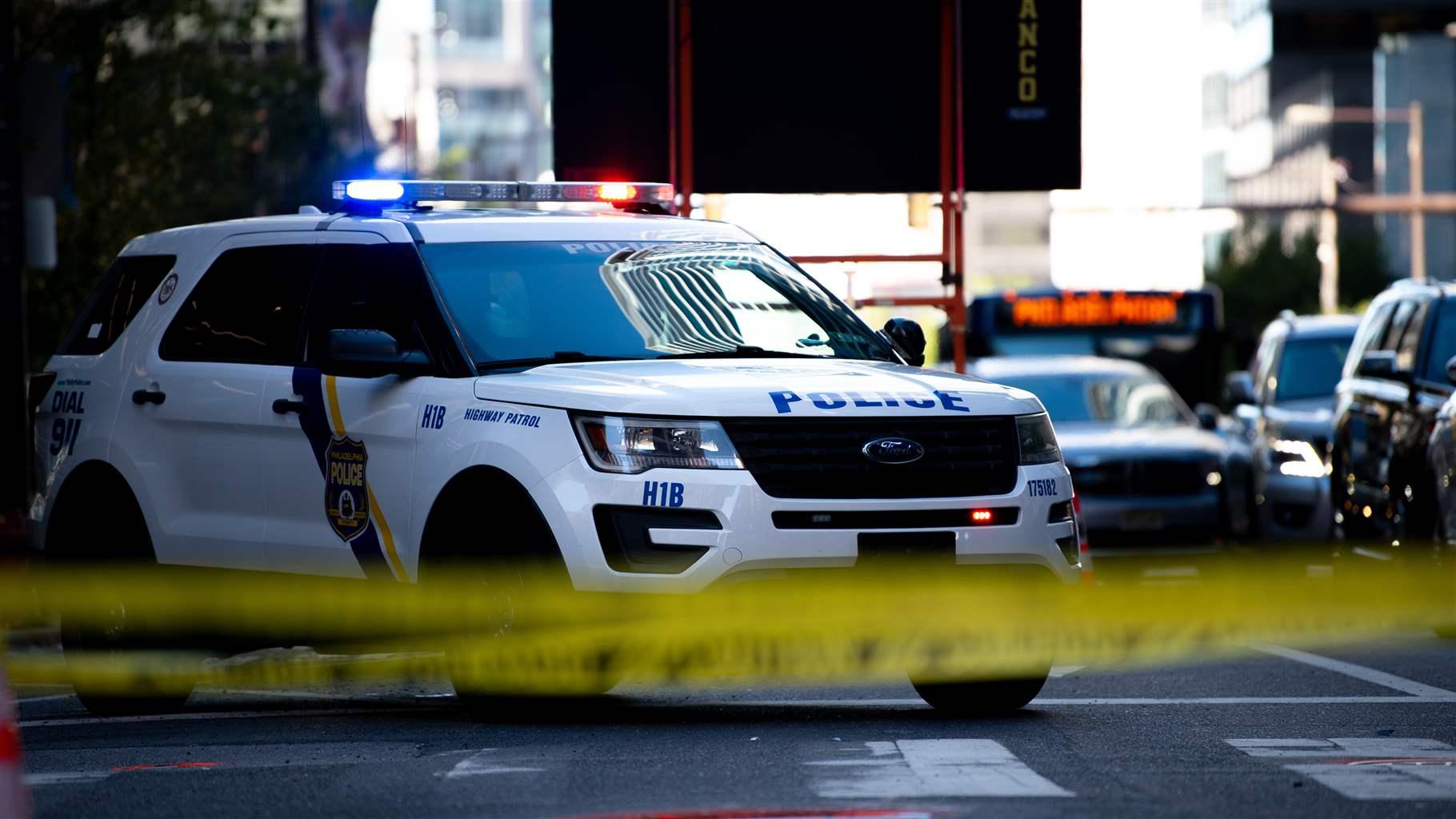 Yellow crime scene tape is blurred in the foreground with a white police SUV in focus behind it. The blue and red lights atop the vehicle are flashing, and the word “Police” appears in blue capital letters across the front of the hood
