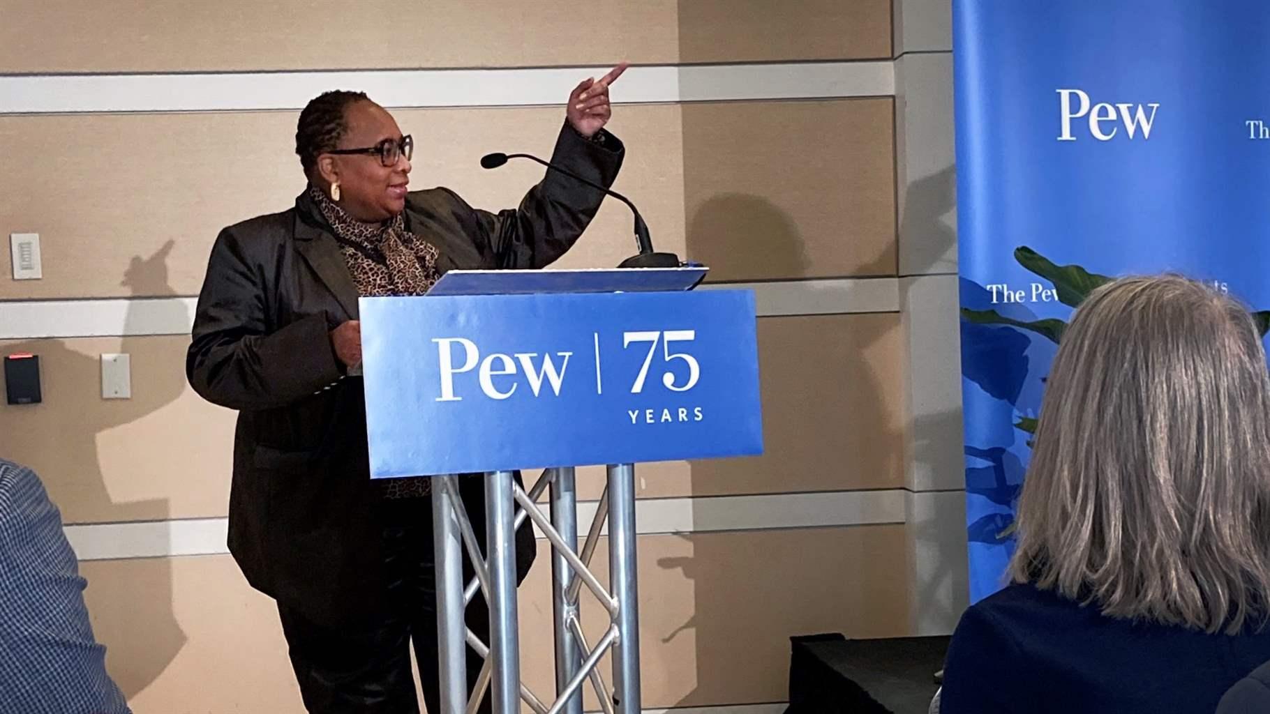 Donna Frisby-Greenwood, wearing a dark pantsuit and a print blouse with a scarf, stands behind a podium with a blue sign in front that reads, in white text: “Pew | 75 years.”