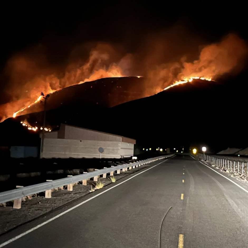 Flames from a raging wildfire rise from hills and create an orange glow in the night sky. In the foreground, a two-lane road, with a guardrail on one side, runs along flat ground straight toward the burning hills. A one-story industrial building is visible on the left. 