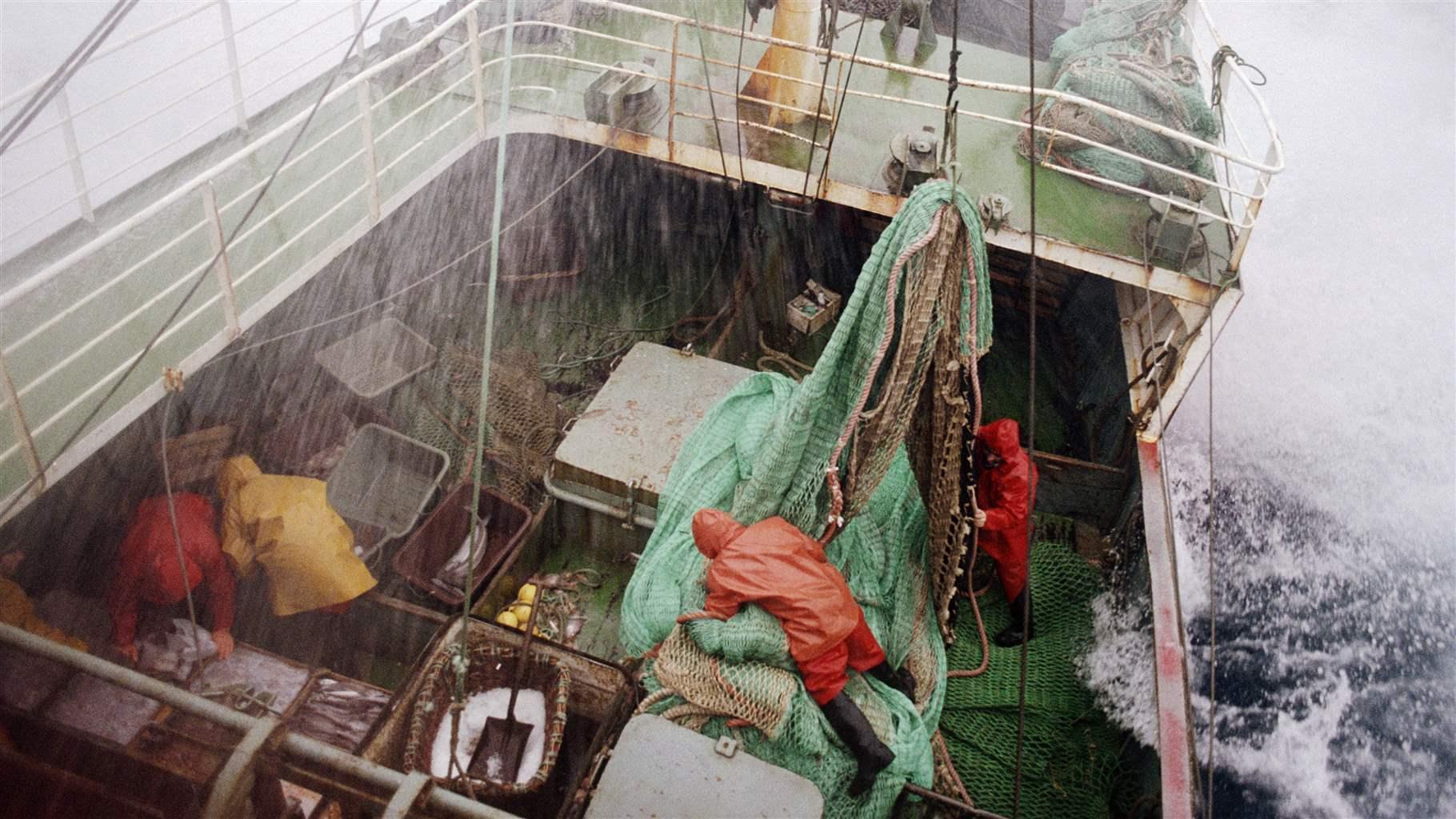 Rain pours down on a fishing vessel at sea, while waves splash the deck. Fishers in orange and red rain jackets work to secure buckets and green fishing nets.