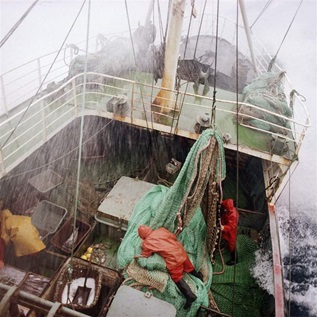 Rain pours down on a fishing vessel at sea, while waves splash the deck. Fishers in orange and red rain jackets work to secure buckets and green fishing nets.