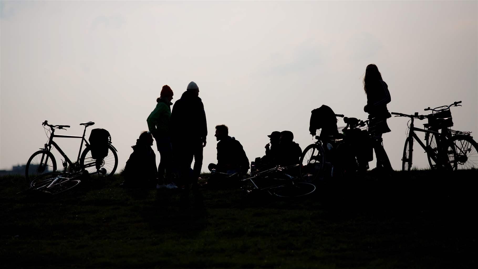 A group of teenagers gather in a park with bikes, their silhouettes in high contrast to the cloudy sky behind them. 