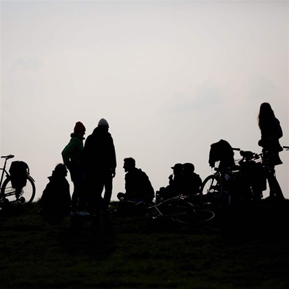 A group of teenagers gather in a park with bikes, their silhouettes in high contrast to the cloudy sky behind them. 