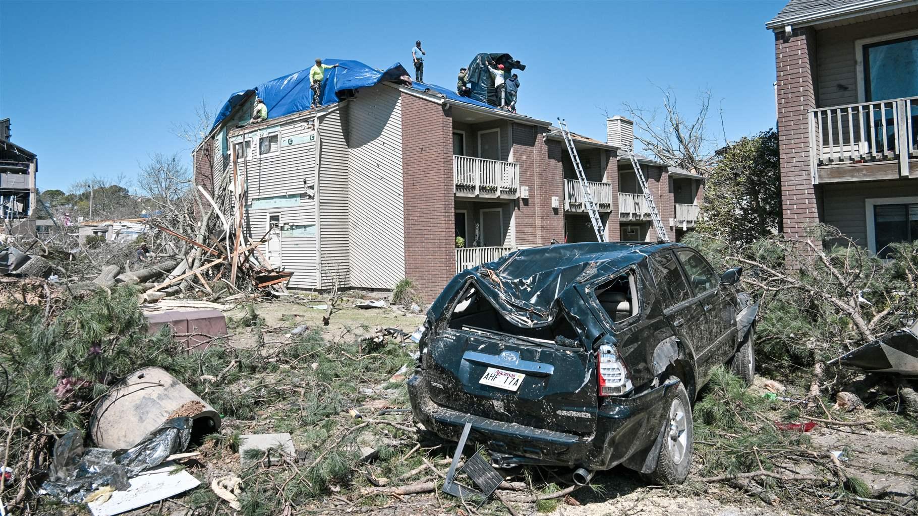 Six workers secure a large blue tarp on the roof of a two-story condominium under a cloudless blue sky. In the foreground is a badly damaged black SUV, along with large branches and pieces of homes—evidence that a strong storm recently struck.   