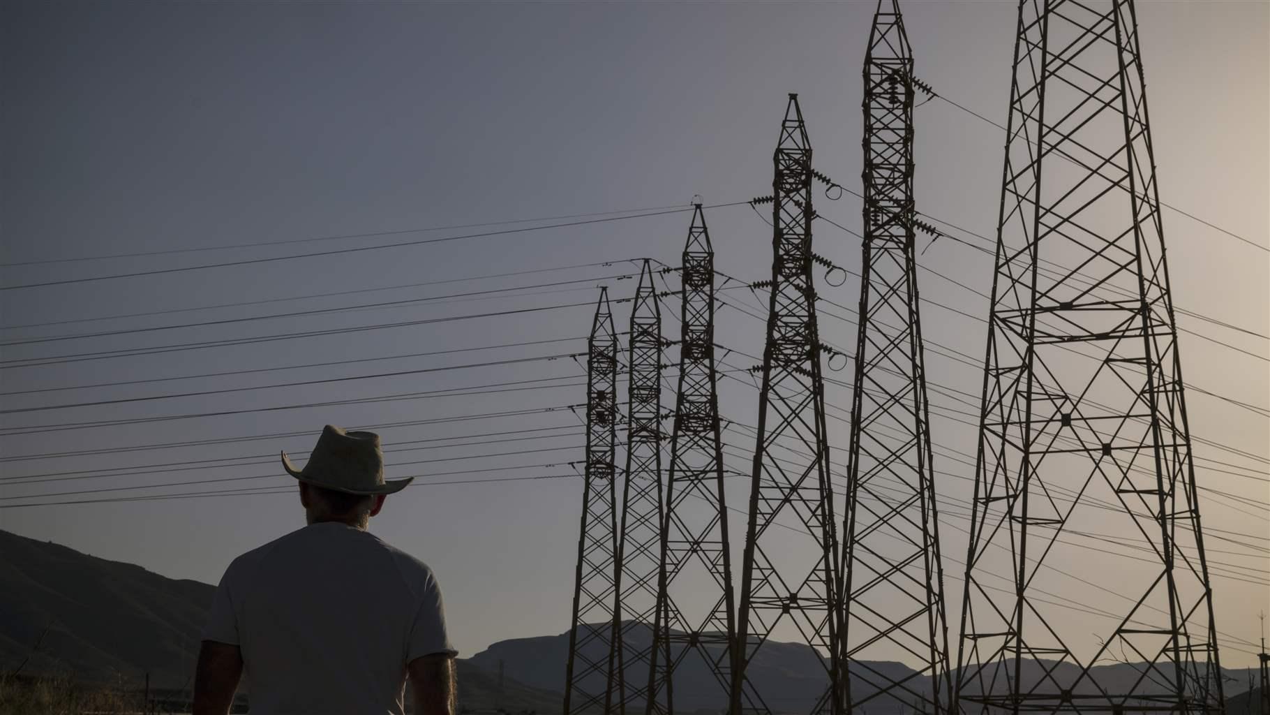 A person cloaked in shadow, wearing a cowboy hat and t-shirt, stands in front of an array of electrical transmission towers. The sky has the pale light of either sunset or dawn.