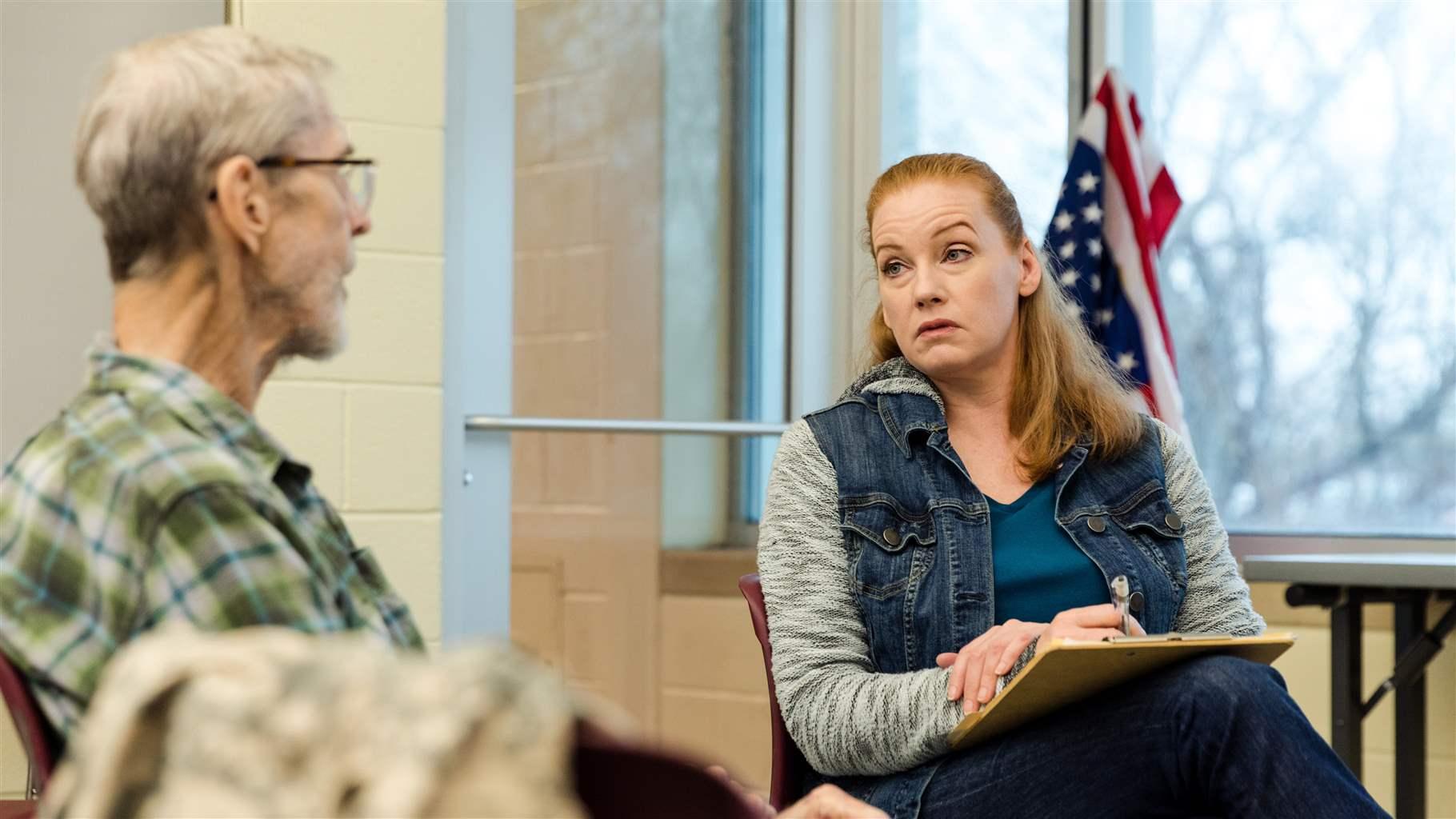 A person with shoulder-length red hair, wearing a gray shirt and denim vest, looks at a person with glasses, short white hair, and a plaid shirt. They sit in a support group setting in front of a window and an American flag.