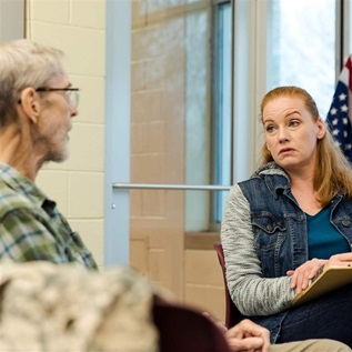 A person with shoulder-length red hair, wearing a gray shirt and denim vest, looks at a person with glasses, short white hair, and a plaid shirt. They sit in a support group setting in front of a window and an American flag.