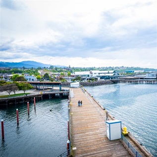 A long and wide wooden pier extends into a large body of water from a shoreline with a mix of low-rise buildings, houses, and trees. Two people are walking on the pier in the distance. Two other piers are visible in the background, as is a faraway mountain.