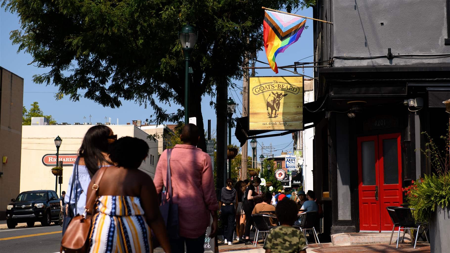 Three people are seen from behind walking along a busy sidewalk on a sunny day. To the right, customers sit at tables under a restaurant sign and a pride flag that’s fluttering slightly.