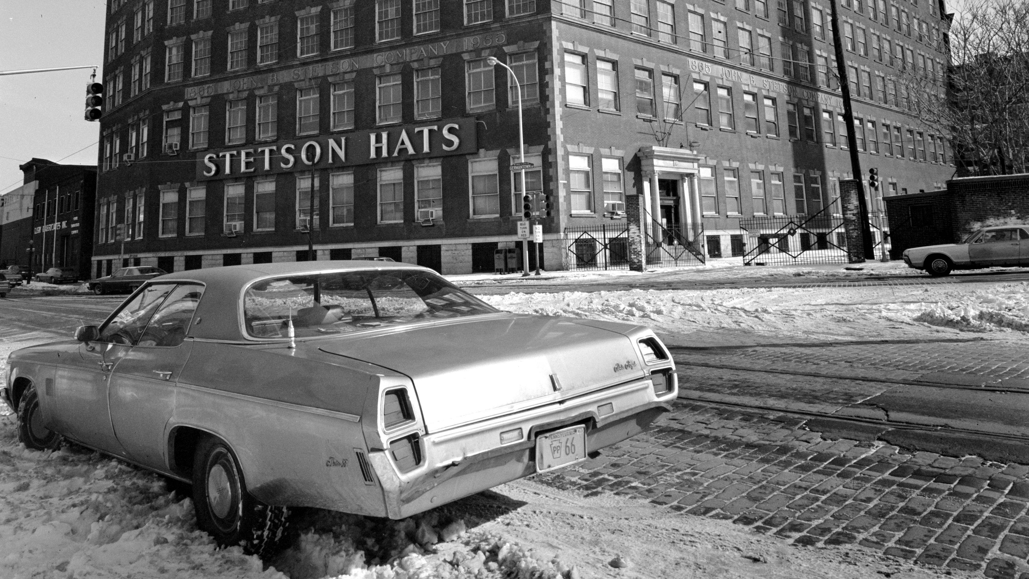 In a black-and-white photo, an eight-story warehouse with many windows and a sign that reads “Stetson Hats” stands on a snowy street. Across the street in the foreground, an older-model car is parked in the snow.