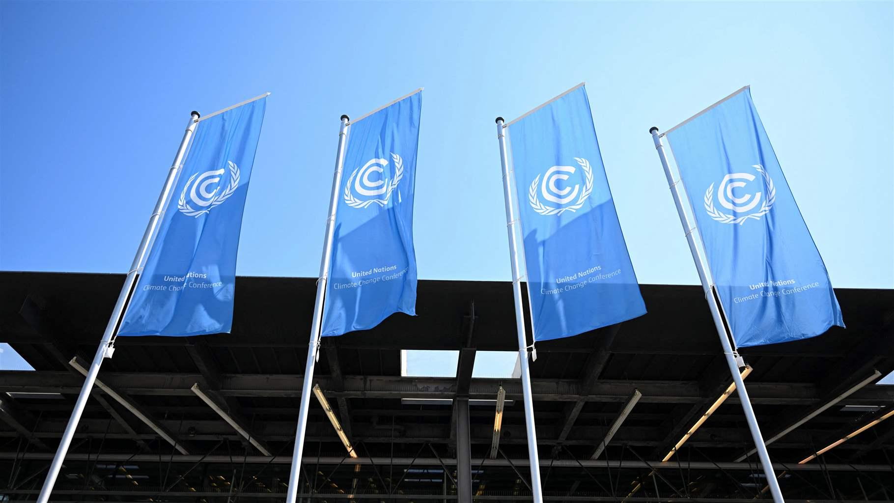 Four identical long, blue, rectangular flags, each with a white United Nations logo and each on its own flagpole, fly in front of a modern conference building. The image is taken from the ground looking up, so only the rook and top couple of floors of the building are visible, in front of a stark blue cloudless sky. 