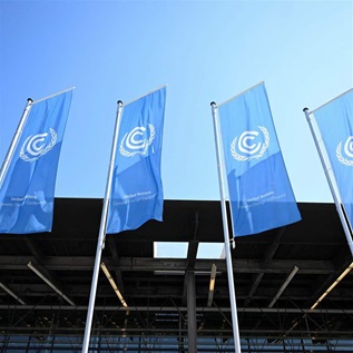 Four identical long, blue, rectangular flags, each with a white United Nations logo and each on its own flagpole, fly in front of a modern conference building. The image is taken from the ground looking up, so only the rook and top couple of floors of the building are visible, in front of a stark blue cloudless sky. 