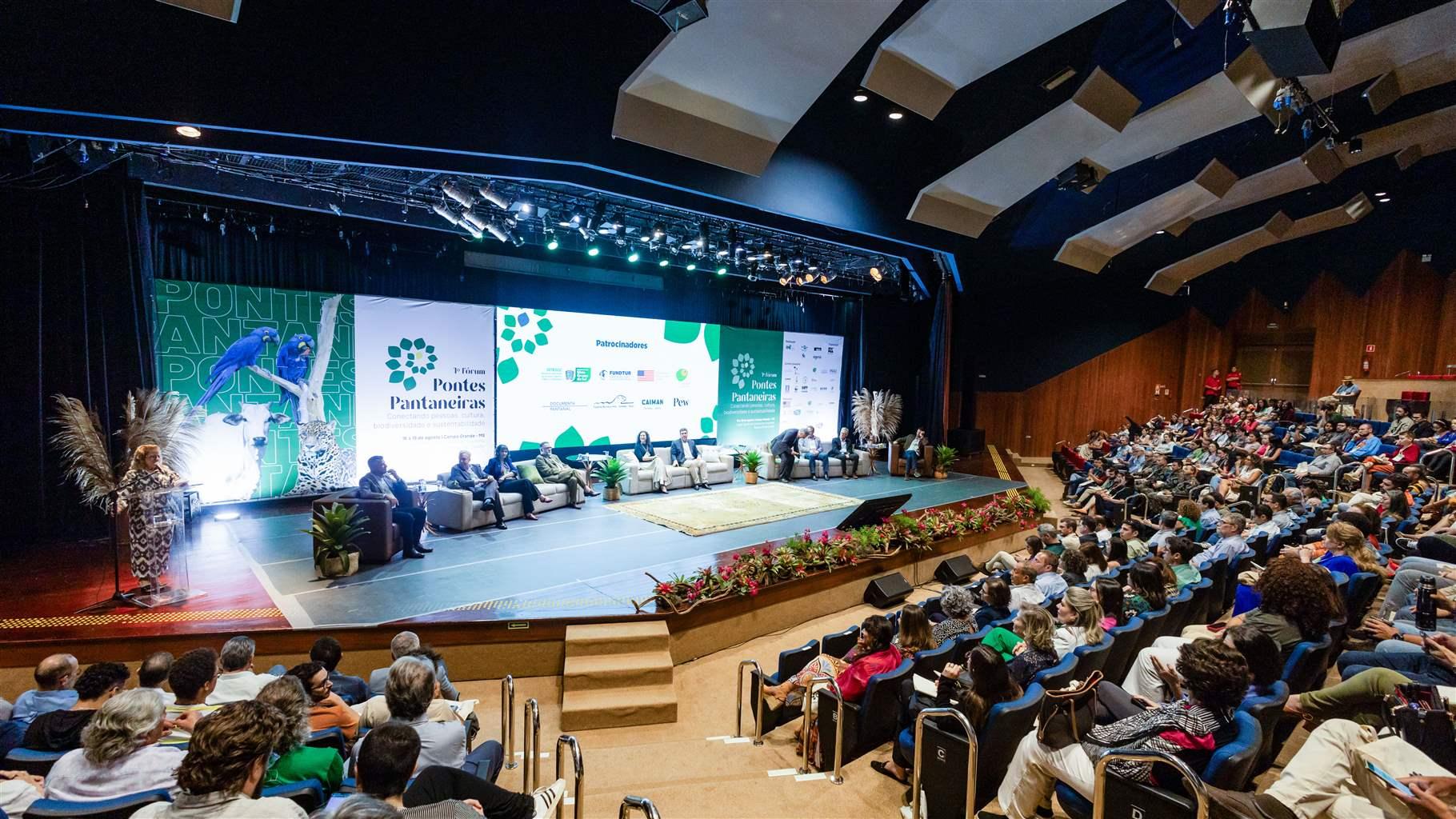 Hundreds of people sit in a large auditorium watching a panel discussion with 10 participants onstage. A large screen behind the panelists features a photo of blue and yellow macaws, a jaguar, and a cow, while others announce the title of the meeting and a list of its sponsors.