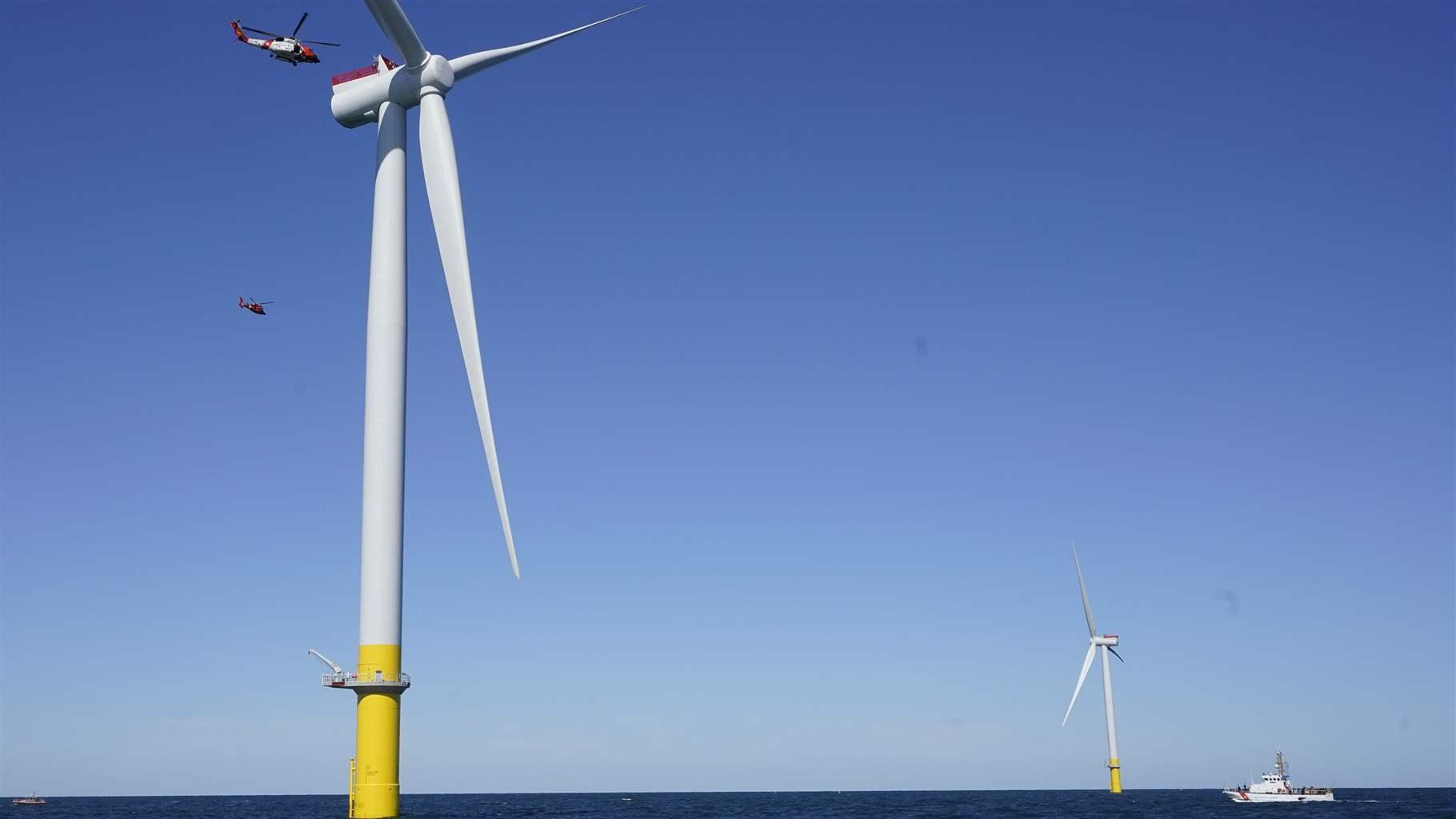 A U.S. Coast Guard helicopter and ship approach two wind turbines in clear ocean waters under sunny blue skies.