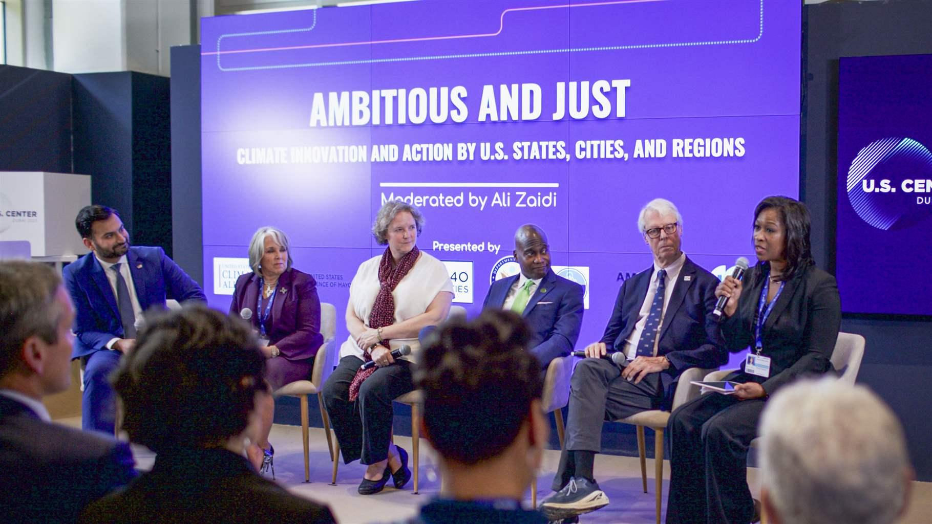 Six people sit on a stage at a panel discussion at an event, with five of them looking on as the person at the far right speaks into a microphone. Behind them is a large banner that says “Ambitious and Just: Climate Innovation and Action by U.S. States, Cities, and Regions.”