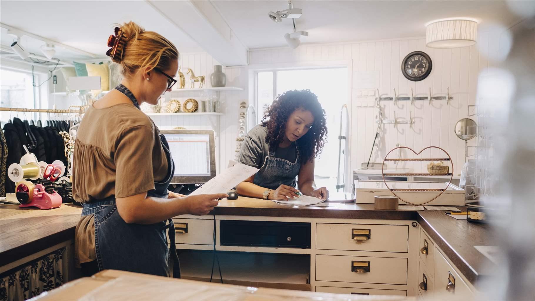 Two women wearing aprons go through paperwork together in a small shop.