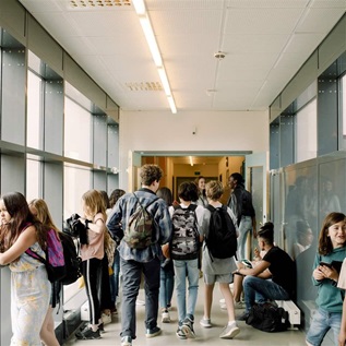 Students gather in a brightly lit hallway between classes, some walking with backpacks and others leaning against green walls, chatting with friends.