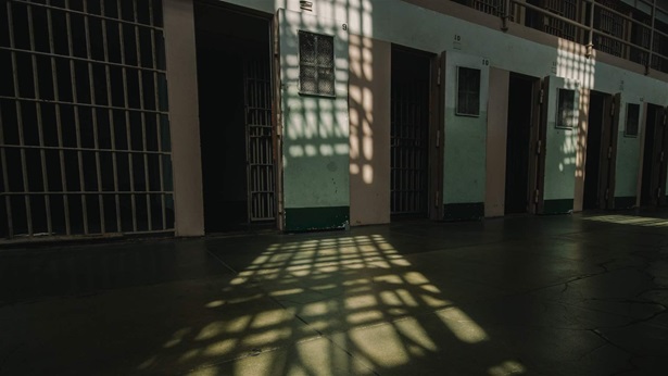 The sun filters through bars in a jail, casting a grid of shadows on the floor in front of the cells. 