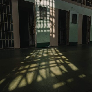 The sun filters through bars in a jail, casting a grid of shadows on the floor in front of the cells. 