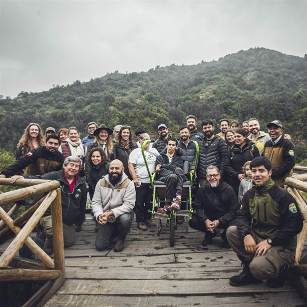 On an overcast day, more than two dozen people pose at a scenic overlook bordered by cactuses, with a green hill as their backdrop. Most are wearing jackets and standing on a wooden platform lined with sturdy wood railings, while some kneel or squat in front. At the center of the group, one person sits in a single-wheeled wheelchair designed to navigate accessible trails like this one.