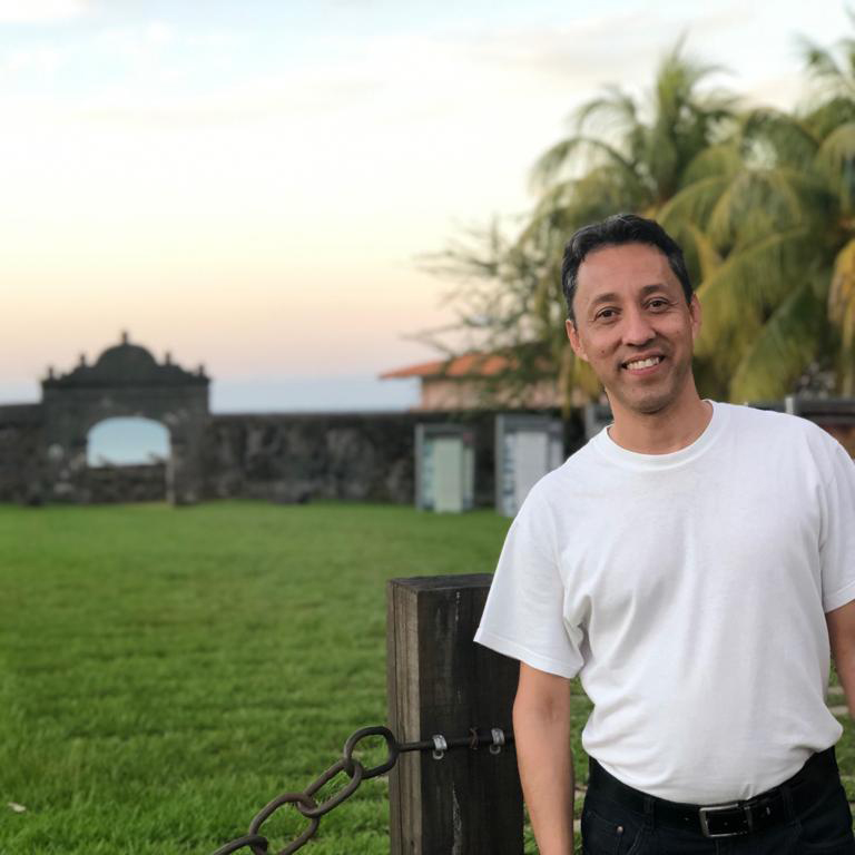 A man in a white T-shirt and dark pants smiles as he stands before a grassy area, with a stone gate and water in the distance.