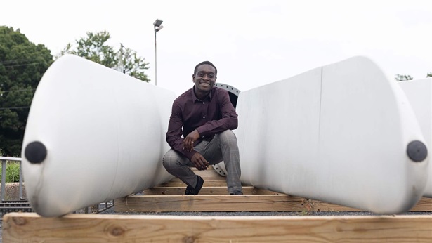 A person in a button-down shirt and slacks has a broad smile while crouching between two white wind turbine blades laying on graduated shallow wood block steps. 