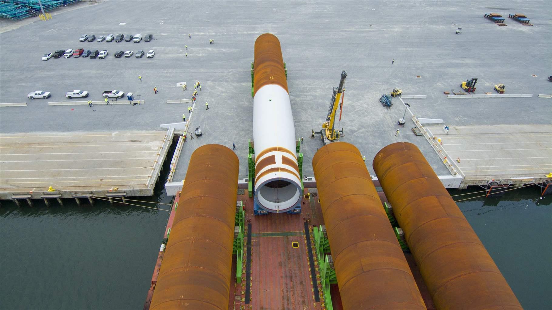An aerial view of a ship port with four massive orange and white tubes partially on the concrete port and partially over the water. A yellow crane is on the shore ready to help take the large structures off the barge.
