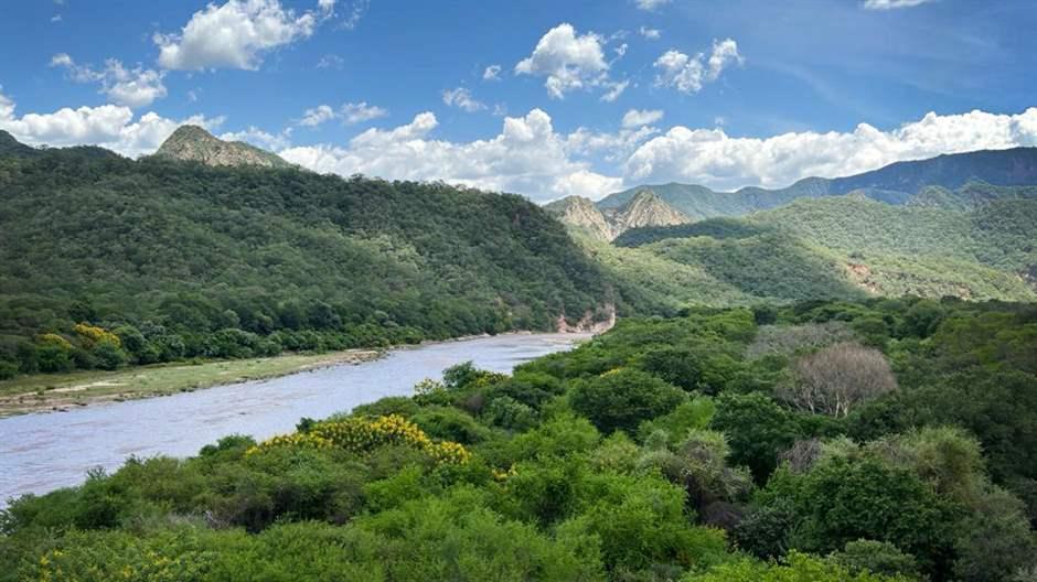 The Parapetí River flows through mountains and valleys in the Bolivian Gran Chaco Forest. Above the river, the blue sky is scattered with white clouds.   