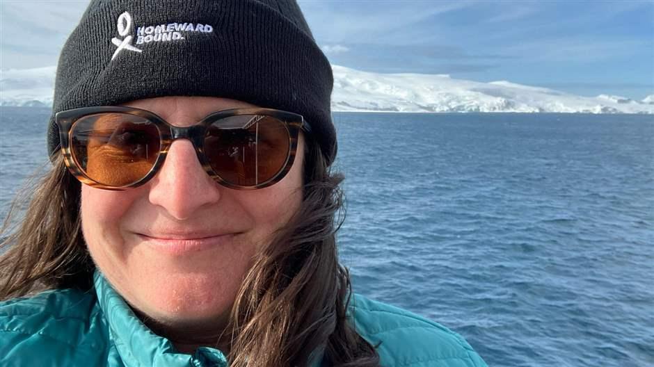 A woman wearing a black beanie, turquoise parka and sunglasses smiles in a selfie. Behind her are ocean waters and white, glaciated terrain in the background. 