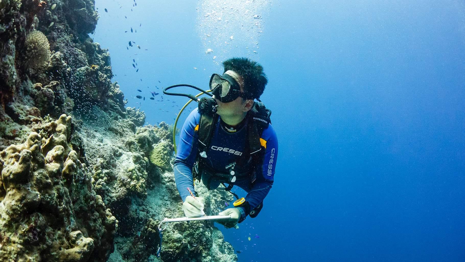 A man wearing scuba gear glances at an underwater coral reef while taking notes on a clipboard.
