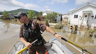 A man in a baseball cap and brown short-sleeved uniform rows a silver boat through muddy floodwaters along a residential street. 