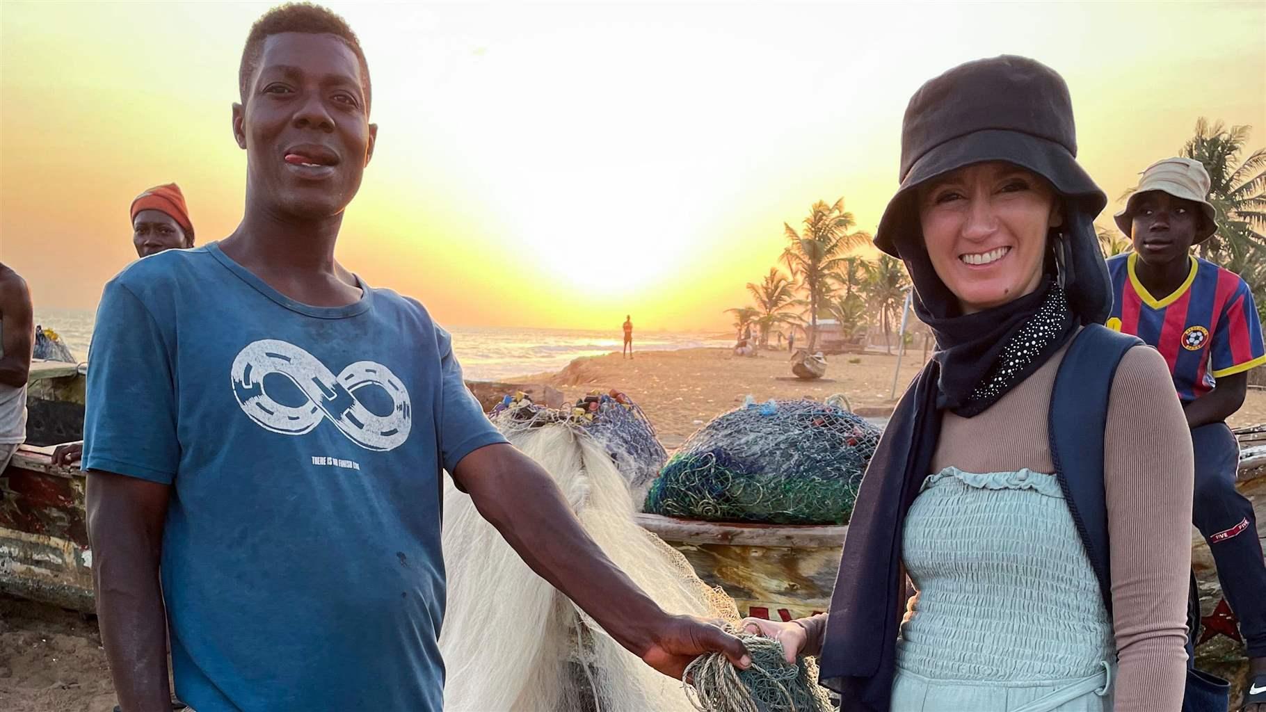 A man and a woman hold a white-and-blue fishing net while standing on a sandy beach as the sun sets behind them. Other individuals stand behind them next to another pile of fishing nets.