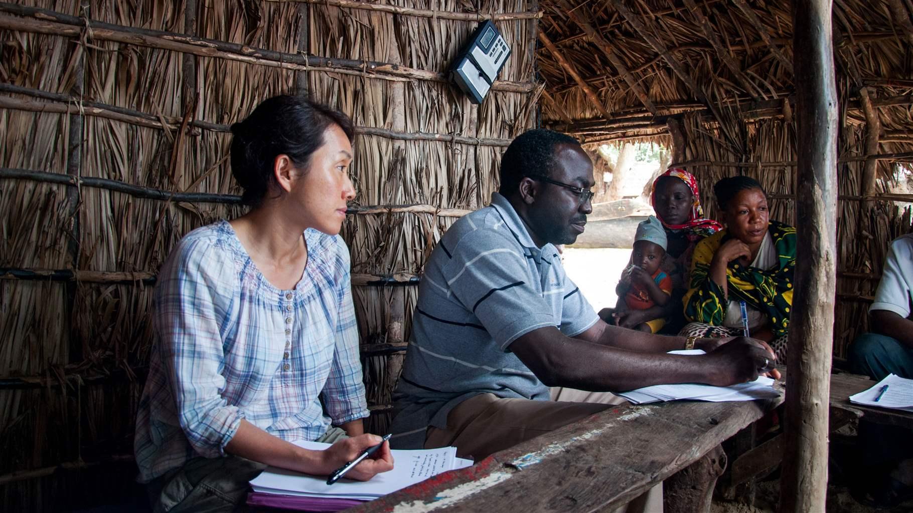 A woman and a man take notes while sitting inside a wooden hut, with a few other people and a young child seated to the side of them in the room.