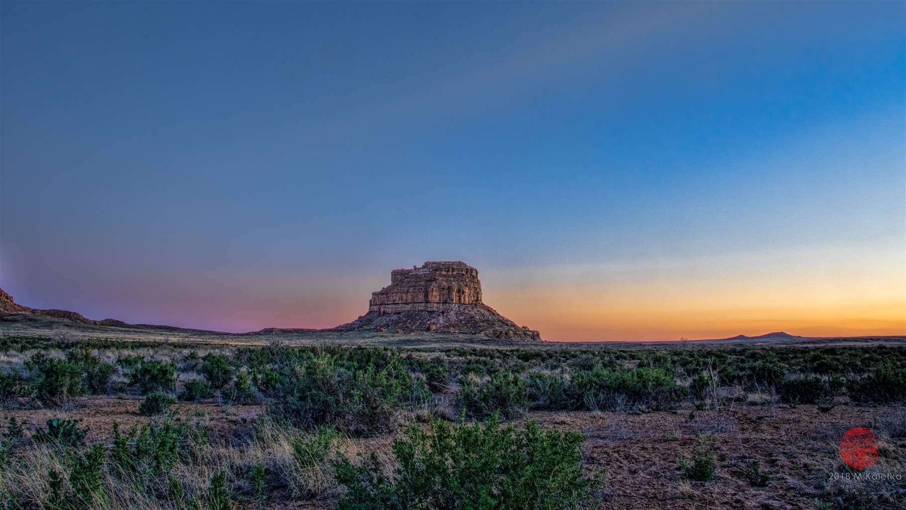 A red-rock butte rises from a desert landscape of low, brittle bushes and sandy dirt. The sky is lit by the colors of a brilliant southwestern sunset.