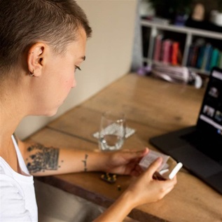 A person holds a box of medication and sits at table in front of a computer during a telemedicine call. 