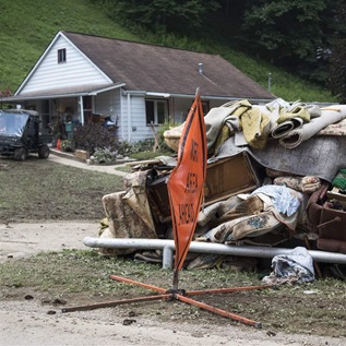 Water-logged furniture and rugs are piled on grass in front of a small white house. Behind the house is a steep grassy hill, and beside the house is a black all-terrain vehicle. In front of the pile is an orange construction sign. 
